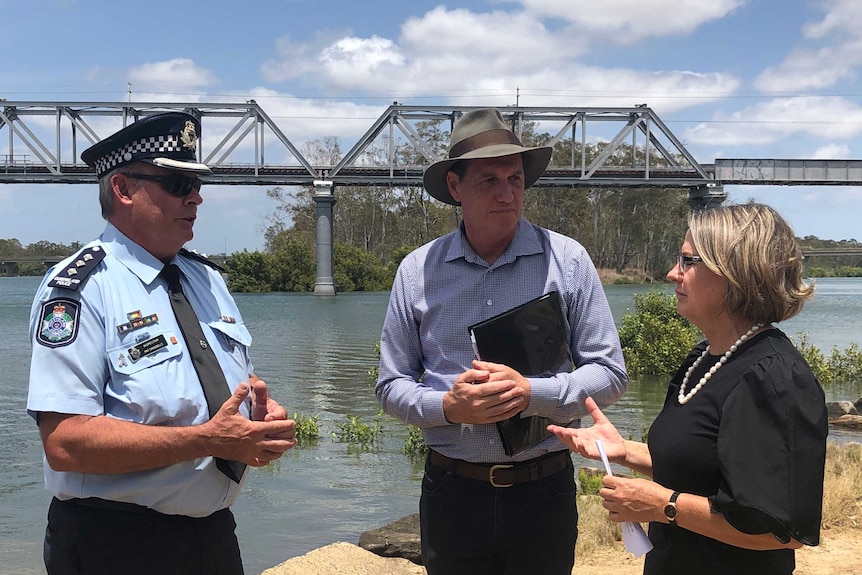 Three people stand talking next to the Burnett River in Bundaberg north with the rail bridge in the background.