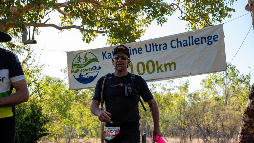 A man in a cap, sunglasses and black T-shirt crosses a finishing line, with a banner reading Katherine Ultra Challenge above him