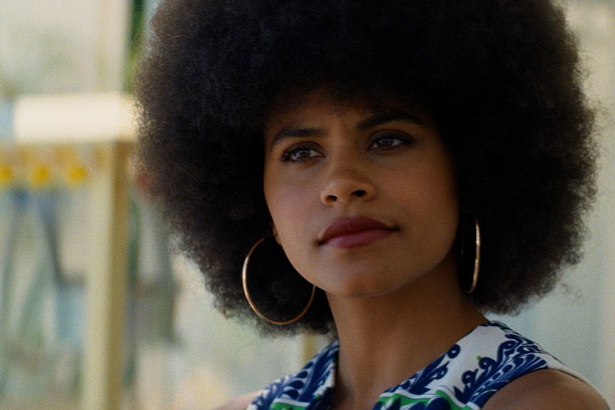 A woman with black afro and serious expression wears large gold hoop earrings and floral top stands in street.