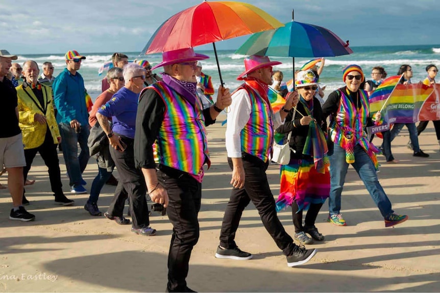 People in brightly-coloured clothes hold rainbow flags as they walk along a beach.