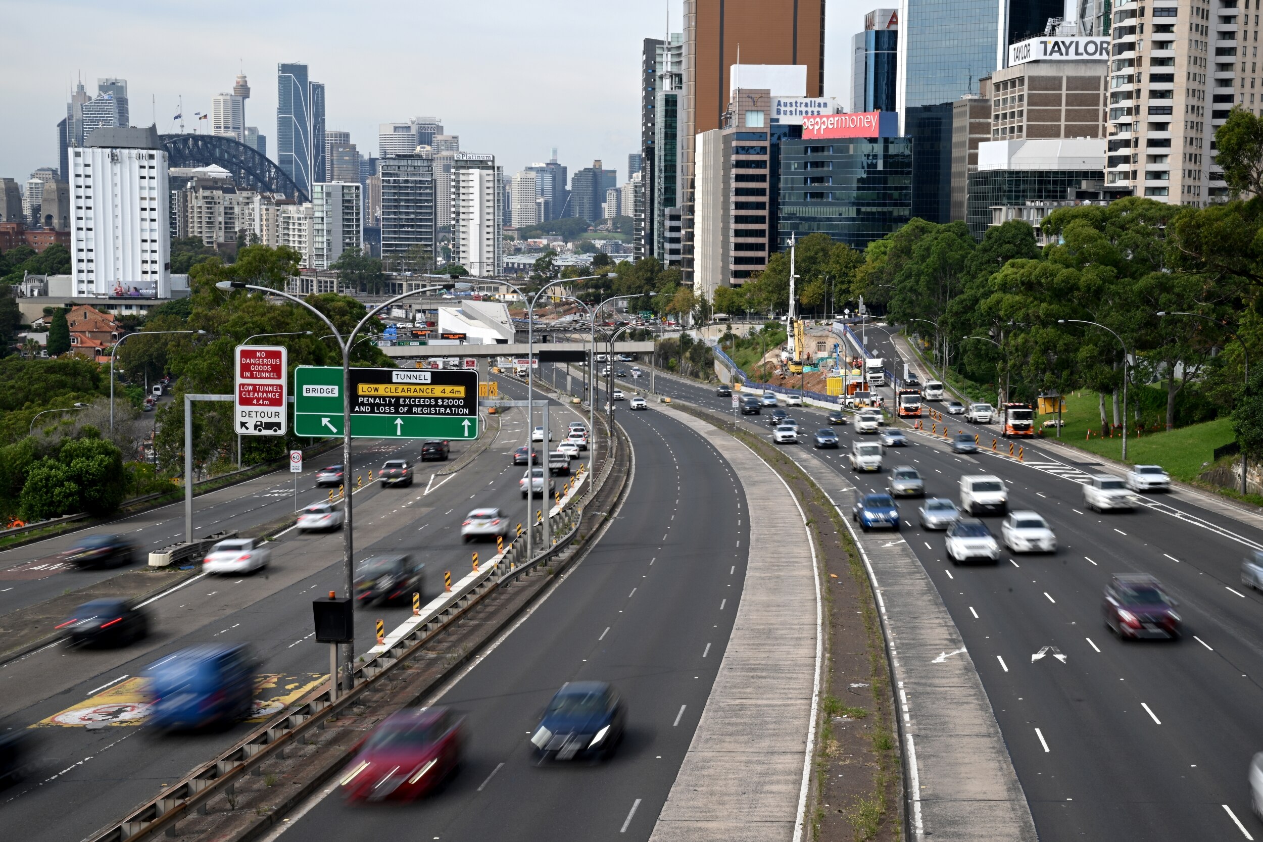 A view of a freeway with multiple lanes and cars taken from an overpass with the Sydney Harbour Bridge and CBD in the distance.