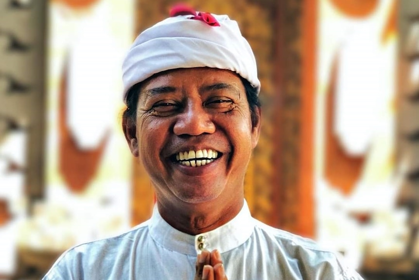 A smiling Balinese man in traditional clothing with his hands pressed together.