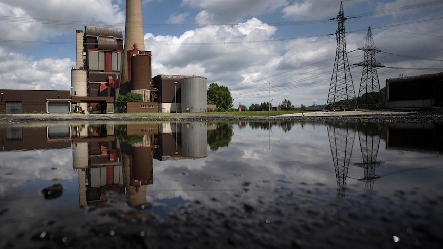 The image of a coal power plant and its chimney stack are reflected in the water of a dam as it stands alongside power pylons.