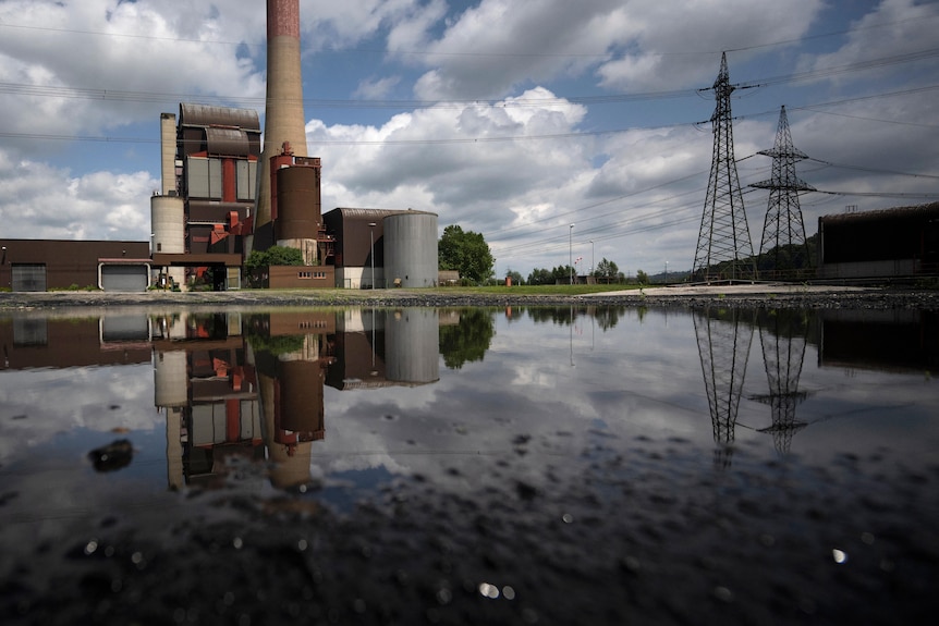 The image of a coal power plant and its chimney stack are reflected in the water of a dam as it stands alongside power pylons.