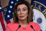 Nancy Pelosi stands at a lectern, with one hand held out in front. There are two microphones