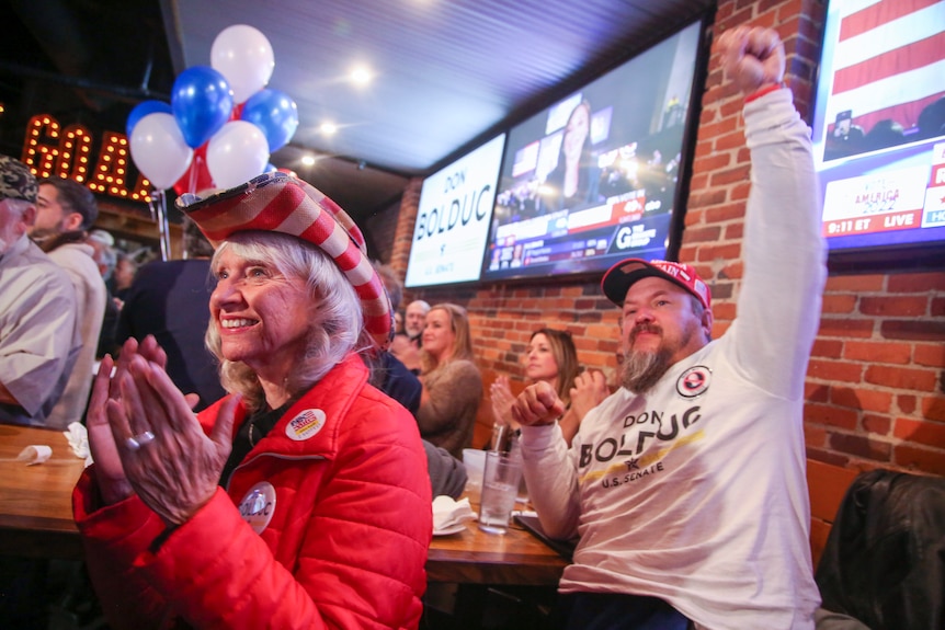 A man raises his arm in celebration as a woman wearing a cowboy hat themed as the American flag applauds inside a venue