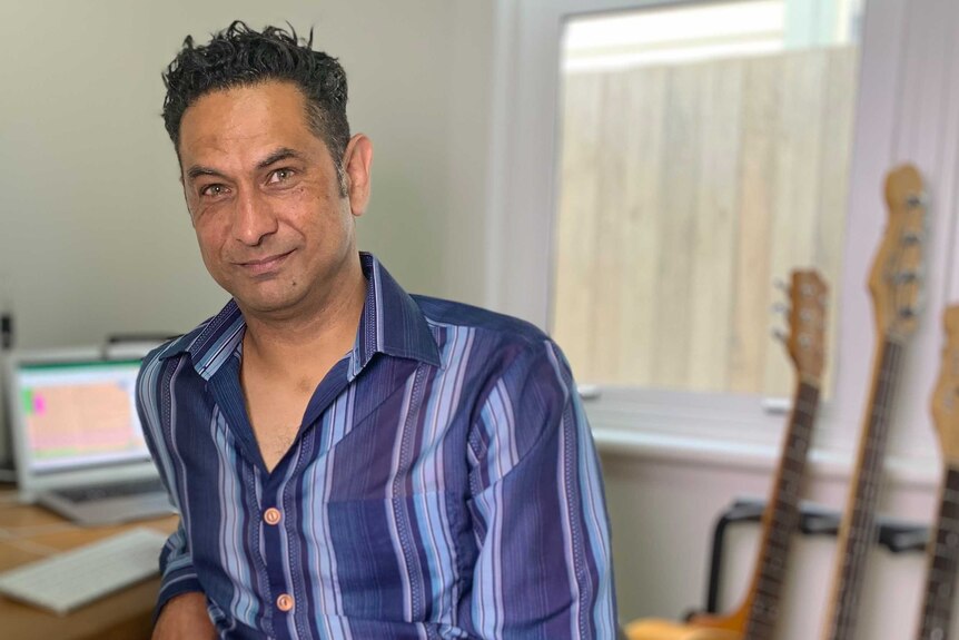 Sean Simmons, wearing a striped blue shirt, in his home office with guitars and a laptop behind him.