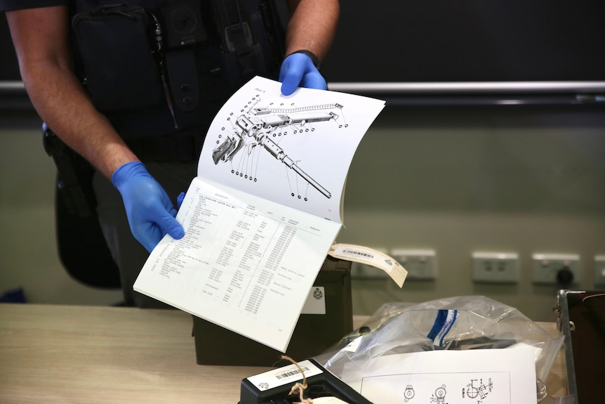 A police officer wears blue gloves while holding a booklet with a black and white diagram of gun components.