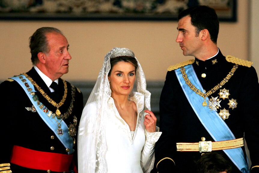 A man dressed in military unform with his bride dressed in a wedding gown stand with a man wearing a blue sash.