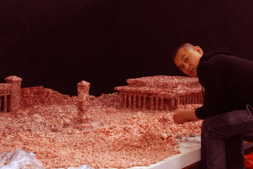 A man sitting next to an artwork presenting the Tiananmen square made out of meat.