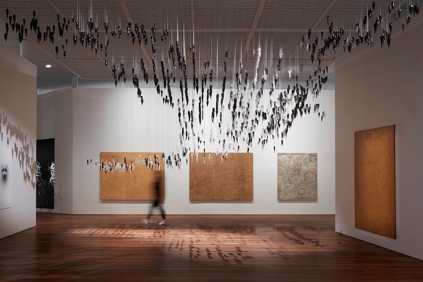 Five Indigenous paintings hang on the walls of a gallery. Dozens of stalactite-like glass pendants hang from the ceiling. 
