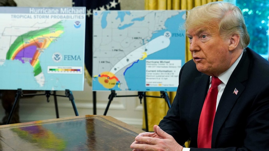 Donald Trump sitting at a desk in front of two maps of the US tracking a hurricane's path.