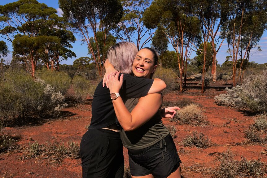 Two women hug and smile in red, sandy landscape with gum trees