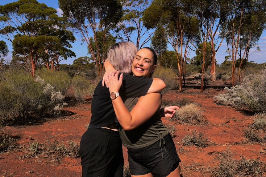 Two women embrace and smile in a red, sandy landscape with gum trees