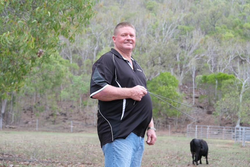 Stephen McAndrew in a black polo shirt holds two thin metal rods. A black cow is in the background.