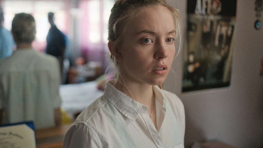 Sydney Sweeney, a blonde white woman with blue eyes, wears a white shirt and stands in a bedroom facing two obscured figures.