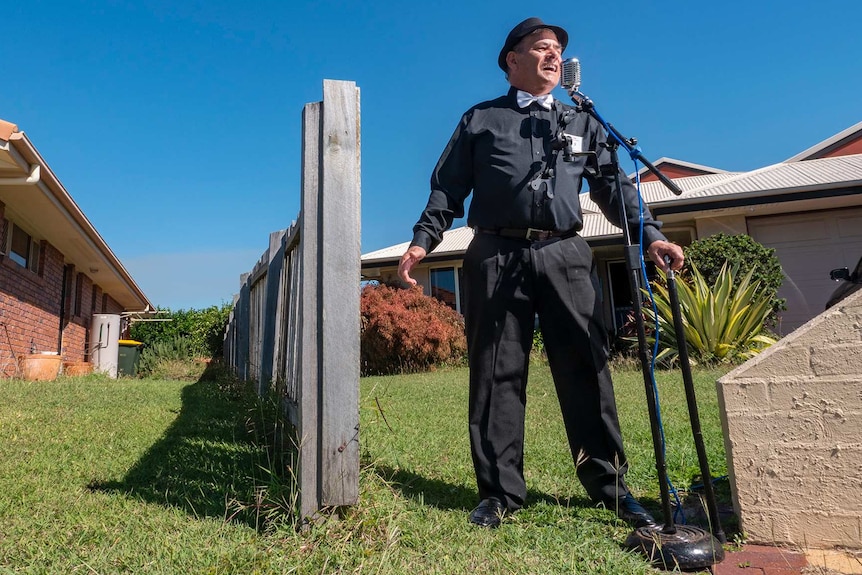 A man standsat the end of his driveway dressed all in black with a silver bow tie singing into an elvis-style microphone.