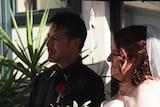A man wearing a black shirt and tie and a woman wearing a white wedding gown stand together