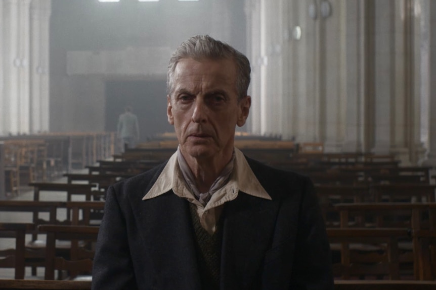 A close-up of a middle-aged man in a suit jacket and scarf sitting looking distressed in a church pew 