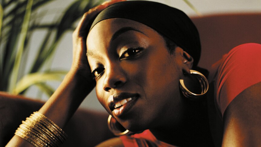 UK rapper Estelle leans her head on her right hand as she smiles at the camera. She wears a black hair wrap.