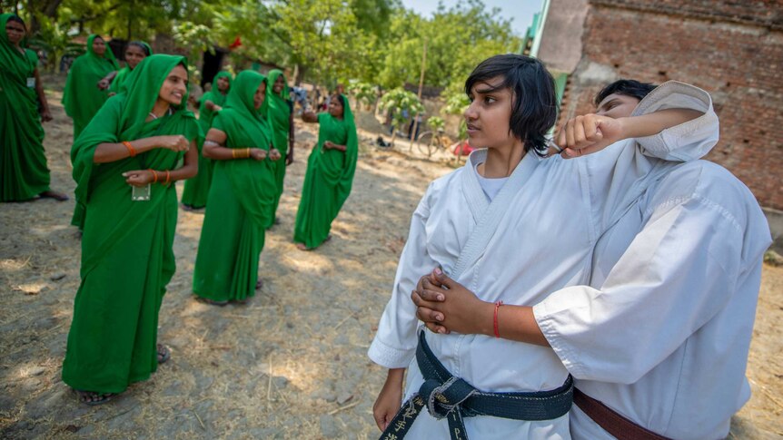 Two young men in karate uniforms demonstrate their moves to women in green saris.