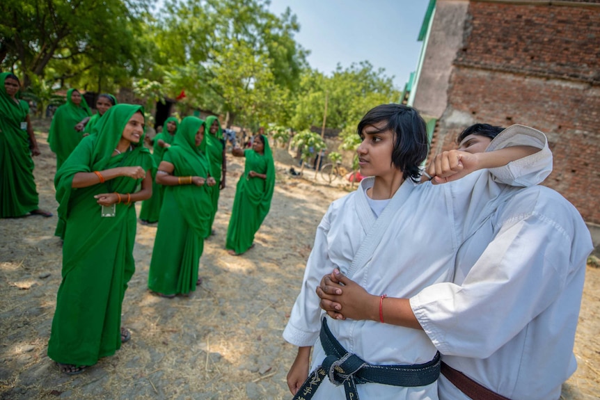 Two young men in karate uniforms demonstrate their moves to women in green saris.