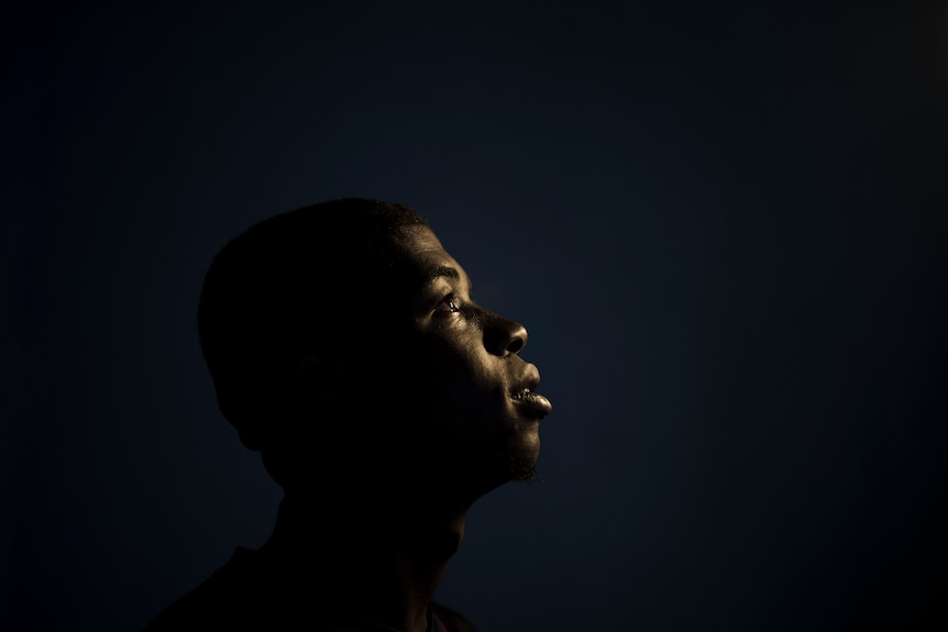 A silhouette photo of a young, black man in a dark room, his face tilted upwards and is bathed by a light source.