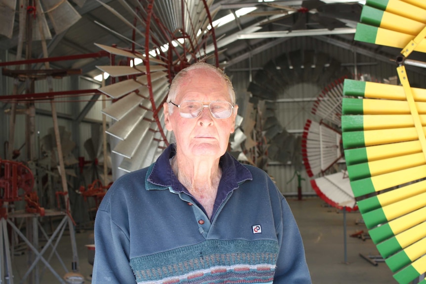 An elderly man stands in front of several large sets of windmill blades within a large tin shed.