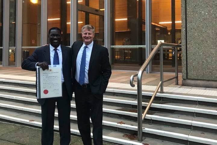 Two men standing in front of Sydney's law courts. William Orule holds a certificate.
