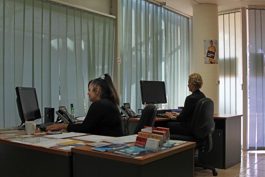 The interior of an office with some afternoon light coming through blinds. Two women sit at computers.