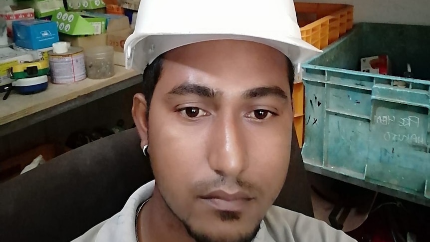 A young South Asian man with a mournful expression wearing a white construction helmet sits in workshop.