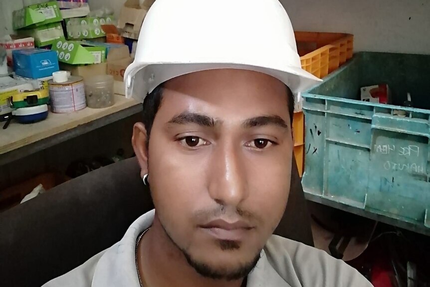 A young South Asian man with a mournful expression wearing a white construction helmet sits in workshop.