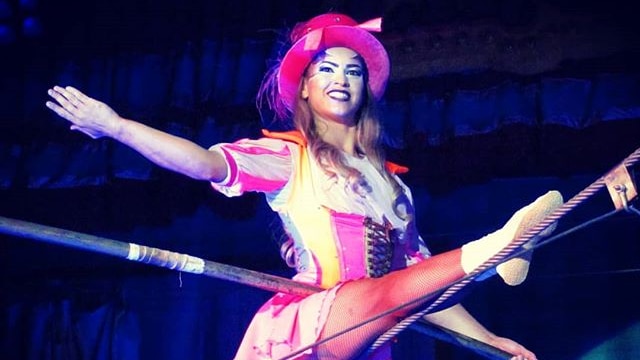 A an aerial performer in a pink hat doing the splits on a wire.