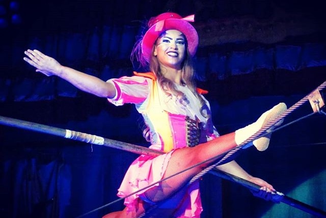 A an aerial performer in a pink hat doing the splits on a wire.