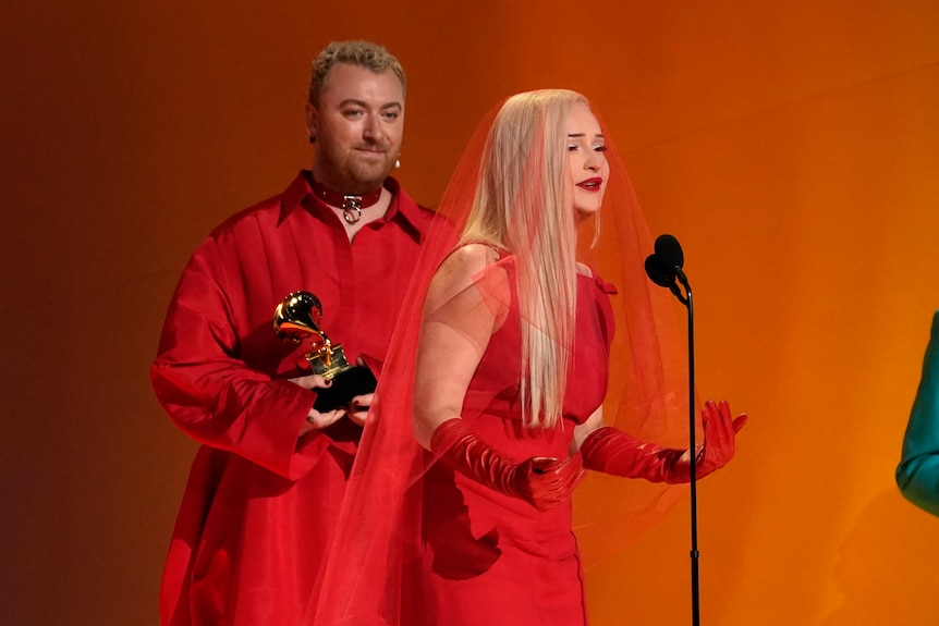 A blonde woman in a bright red, veiled dress speaks into a microphone as Sam Smith stands behind her holding a Grammy trophy.