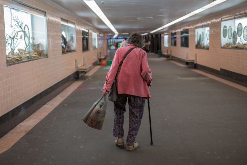 An elderly lady uses a walking stick to make her way through a train tunnel