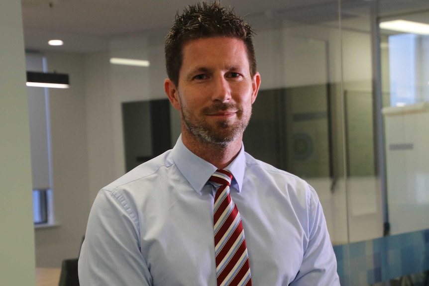 A man in a shirt and red striped tie smiles for the camera in an office.