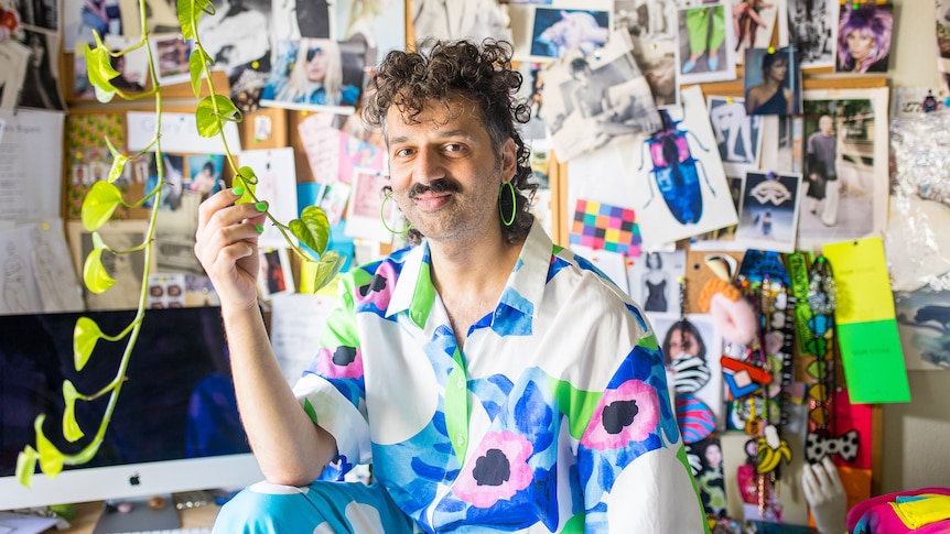 A Maltese Australian man with curly grey hair wears a floral shirt and neon green earrings, in front of a design vision board