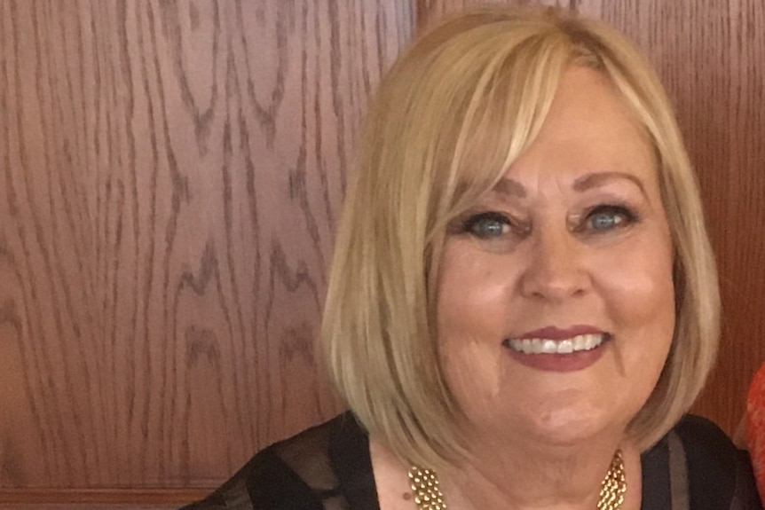 A woman with short blonde hair, gold necklace and black shirt looks ahead and smiles widely.