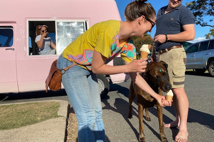 A woman and man with their pet dog, the woman holding an ice cream while she feeds an ice cream to the dog