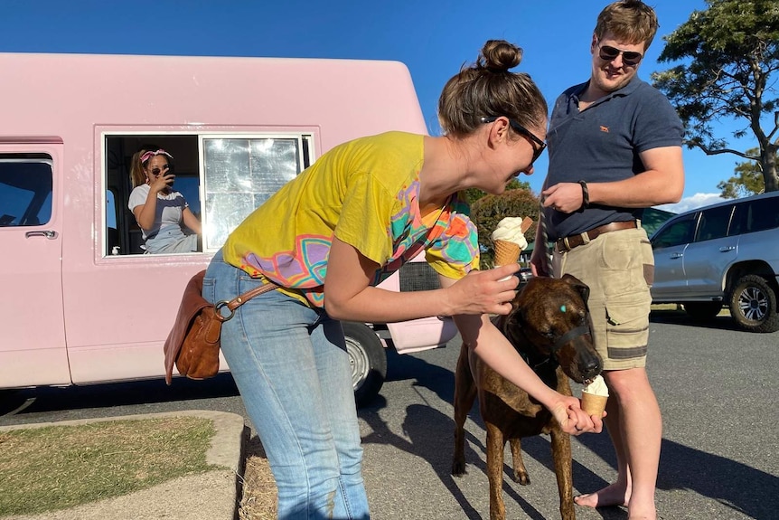 A woman and man with their pet dog, the woman holding an ice cream while she feeds an ice cream to the dog