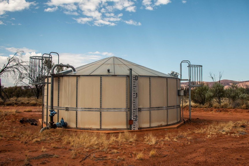 A large tank sits in front of a mountain range on red dirt. A water level meter is attached to the front of it.