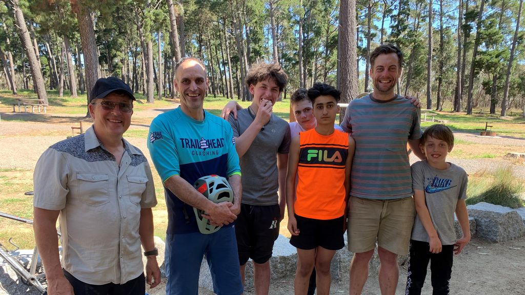 A group of men and boys stand together, one holding a bike helmet, with trees in the background.