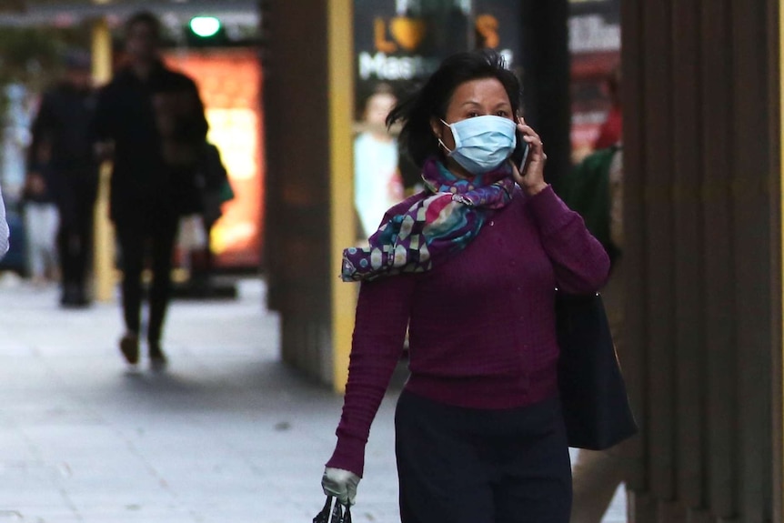 A woman walks along St Georges Terrace speaking on the phone, wearing a mask and carrying a bag