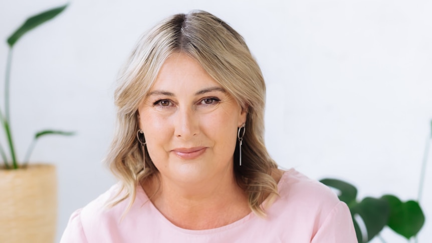 Sonya Lovell wearing a light pink top in front of a white wall with two indoor plants