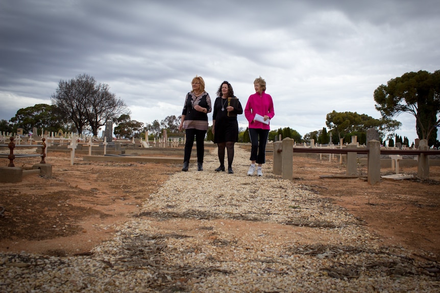 Three women stand on a rock path in the middle of a cemetery.