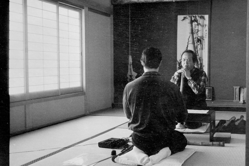 Two men sit across a table on traditional Japanese tatami mats during a shakuhachi lesson.