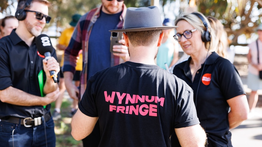 A man wears a shirt with the words "Wynnum Fringe" as two radio hosts with radio microphones and headsets interview him.