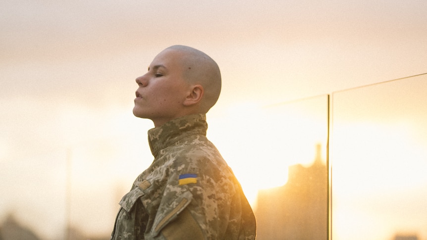 A woman with a shaved head in a soldier's uniform, side profile. 
