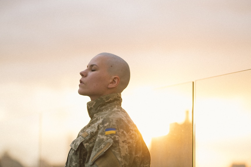 A woman with a shaved head in a soldier's uniform, side profile. 